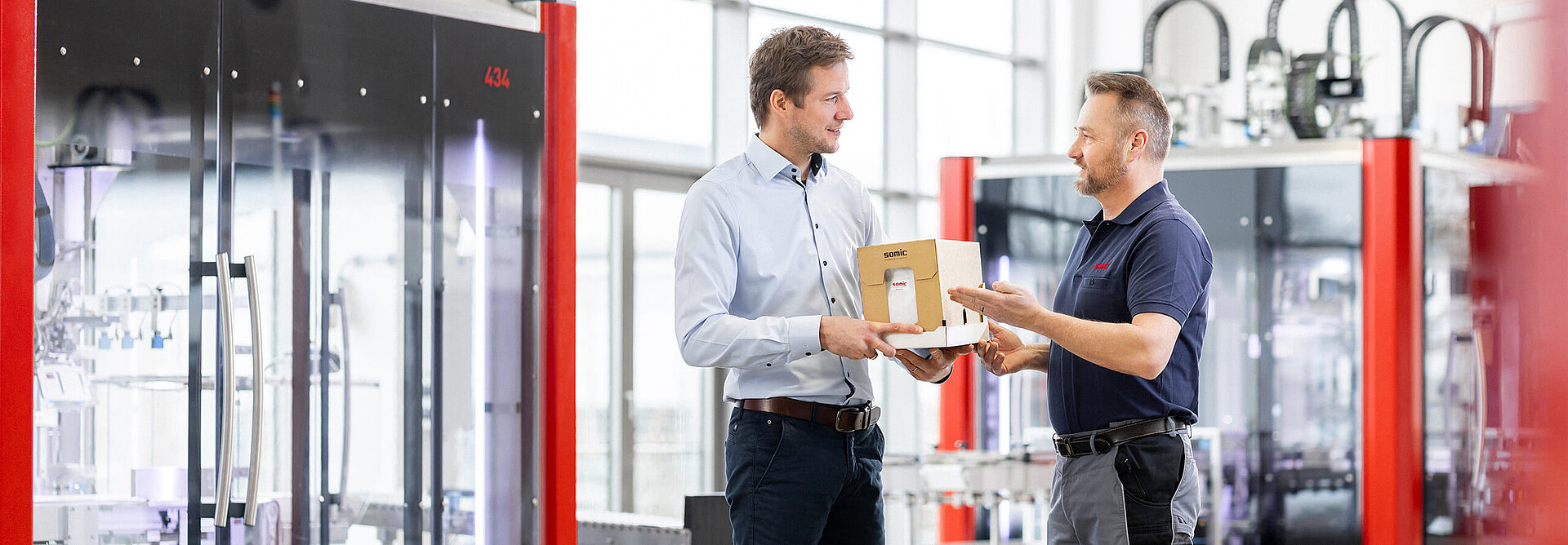 Two employees are inspecting a cardboard blank in front of a SOMIC cardboard packaging machine in the commissioning hall – packaging consulting by SOMIC